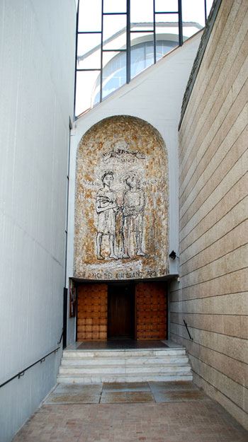 Ingresso ordinario, al termine dell'atrio; nella nicchia sul portale: mosaico con i due santi titolari nellabbraccio di Dio Padre, disegnato da Enrico Castiglioni.